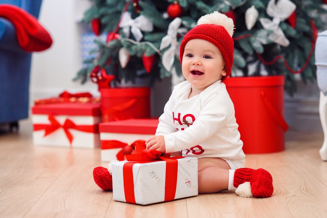Baby wearing red hat and socks sitting with Christmas gift in front of decorated tree for baby first holiday ideas
